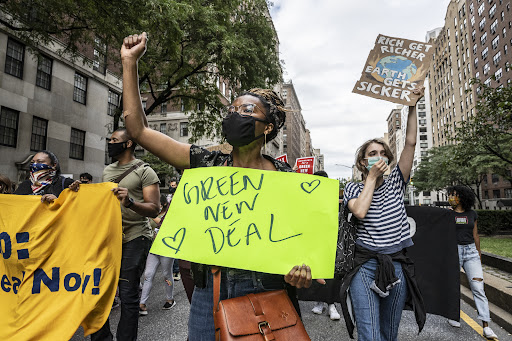 A picture of Dominique at a tax the rich protest, holding a sign that says Green New Deal, raising her right fist in the air, and wearing a covid mask, with other people in the background, one holding a sign that says Rich Get Richer Earth Gets Sicker.